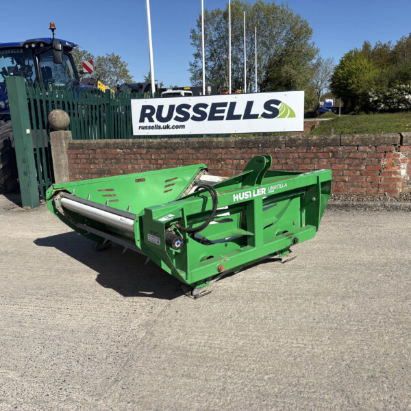 Blue tractor and a green farming implement parked behind a brick wall with a Russell’s sign in a yard under a clear blue sky.