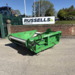 Blue tractor beside a large green harvesting attachment, with a Russell's sign on a brick wall in the background.