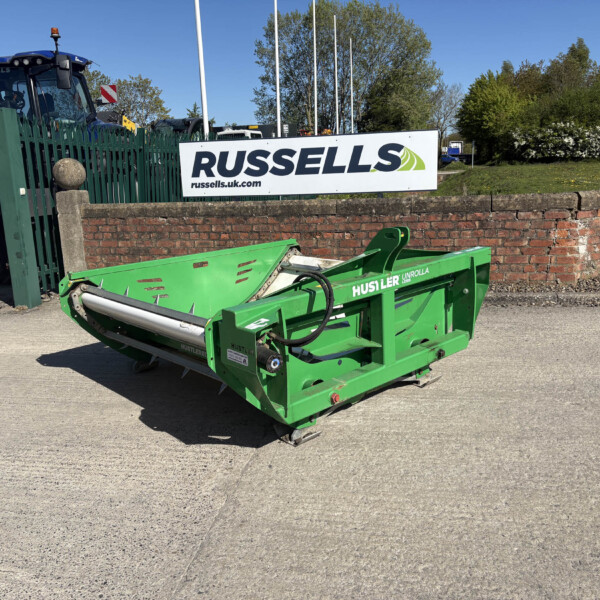Blue tractor beside a large green harvesting attachment, with a Russell's sign on a brick wall in the background.