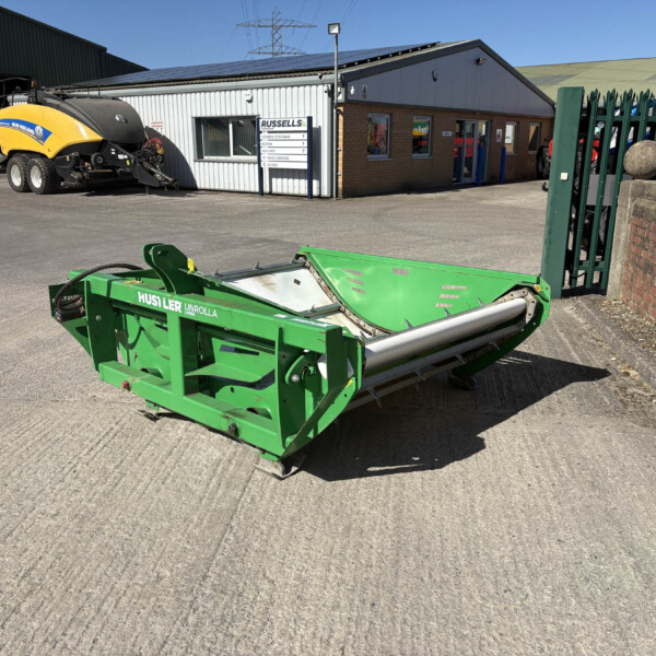 Green agricultural machine in the foreground at an industrial yard, with a yellow New Holland machine and a Russell's building in the background on a sunny day.