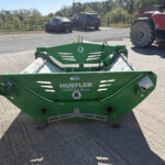 Green Hustler Unroller LX105 farming implement on a trailer parked on a rural road; a red tractor is visible to the right and cars in the background.
