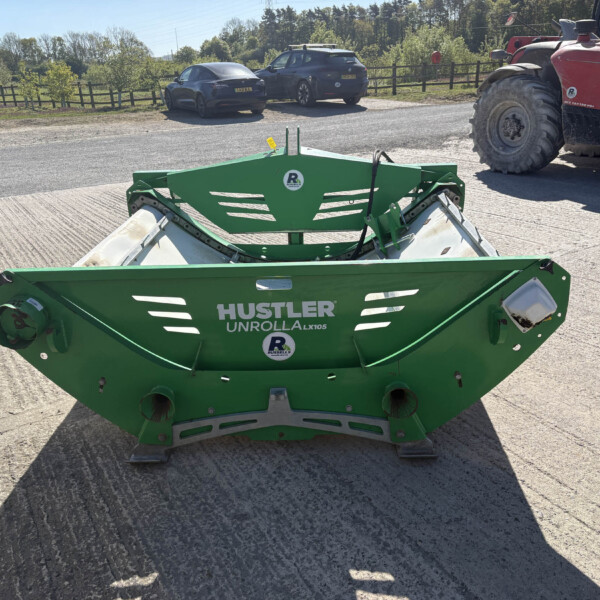 Green Hustler Unroller LX105 farming implement on a trailer parked on a rural road; a red tractor is visible to the right and cars in the background.