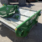 Green agricultural mower/blower attachment with metal spikes and a curved chasis, sitting on dirt beside a tractor; shows warning sticker and brand labels.