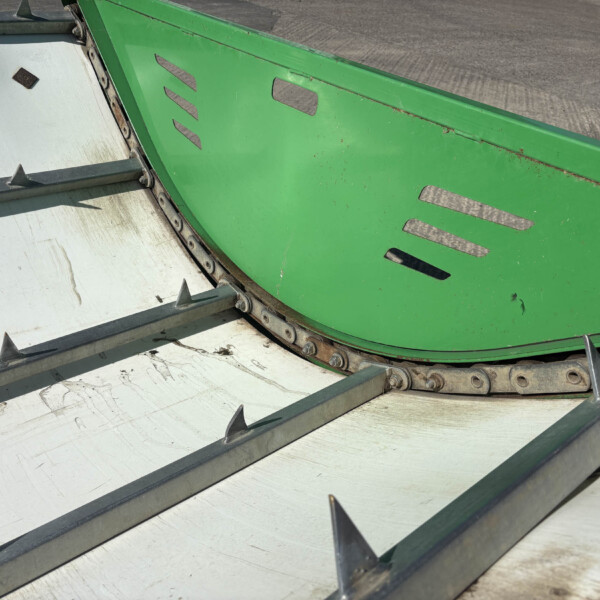 Close-up of green industrial machinery with slotted panels and sharp metal fins along a curved track, outdoors on concrete ground.