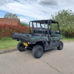 Rear three-quarter view of a Kawasaki diesel 4x4 utility vehicle parked on a residential street, with red hedges and blue sky in the background