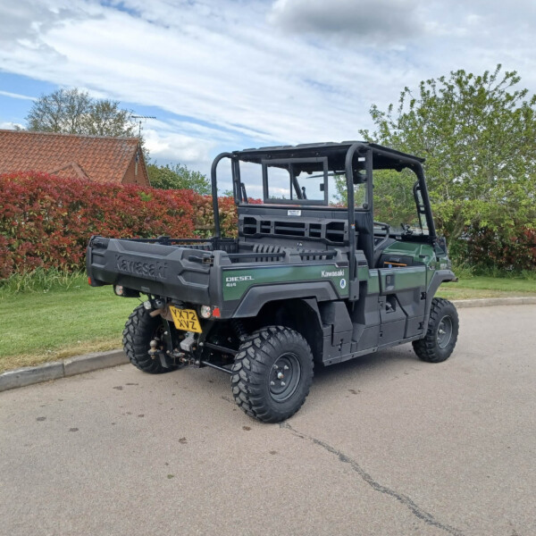 Rear three-quarter view of a Kawasaki diesel 4x4 utility vehicle parked on a residential street, with red hedges and blue sky in the background