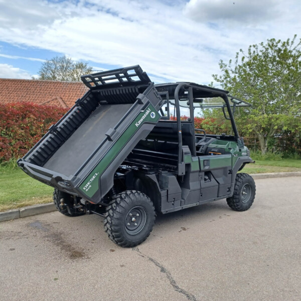 Green Kawasaki dump truck with raised bed dumping load on a suburban street, red-tiled houses and trees in the background.