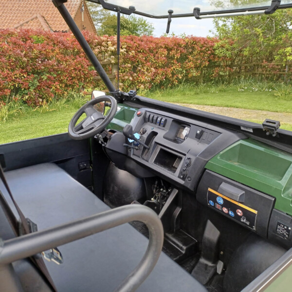 Interior of a green utility vehicle showing steering wheel and dashboard, with red hedges outside the windshield area