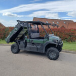 Green off-road utility vehicle with a raised dump bed parked on a street, red hedges and brick houses in the background.