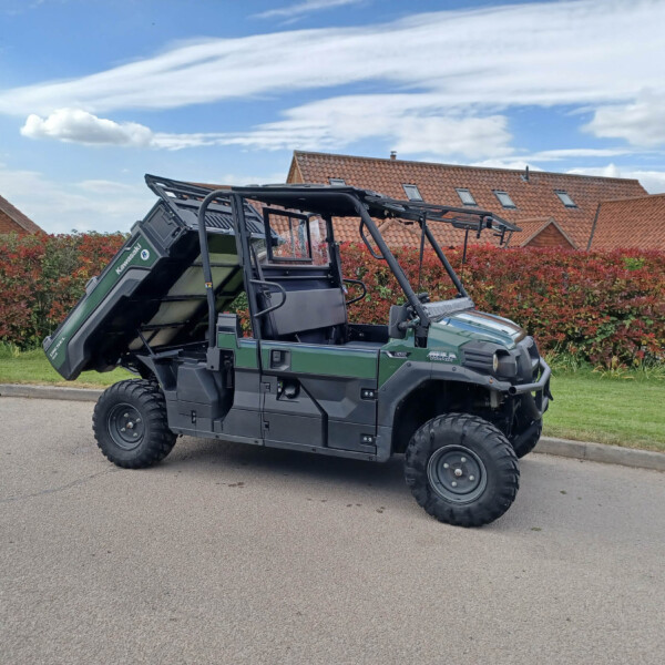 Green off-road utility vehicle with a raised dump bed parked on a street, red hedges and brick houses in the background.