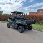 Green Kawasaki diesel 4x4 utility vehicle parked on a suburban street beside a tall red hedge and brick-roofed houses in the background.