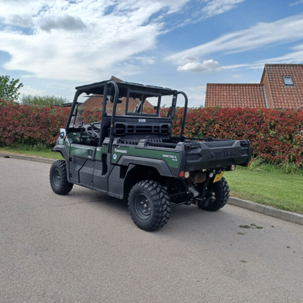 Green Kawasaki diesel 4x4 utility vehicle parked on a suburban street beside a tall red hedge and brick-roofed houses in the background.