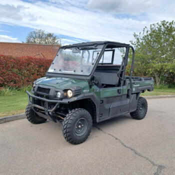 Front three-quarter view of a green Kawasaki UTV with a roll cage, parked in a suburban lot beside red hedges and a cloudy blue sky.