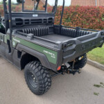 Green Kawasaki Diesel 4x4 utility vehicle with an empty cargo bed, parked near a red hedge and brick houses in the background (rear-three-quarter view).