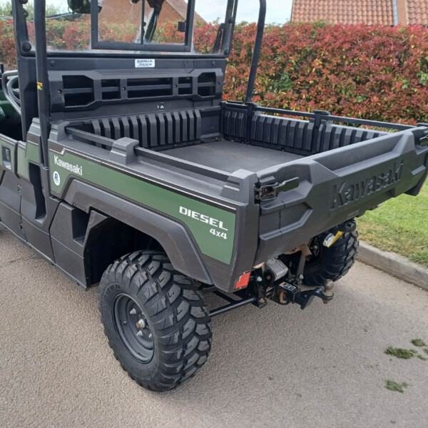 Green Kawasaki Diesel 4x4 utility vehicle with an empty cargo bed, parked near a red hedge and brick houses in the background (rear-three-quarter view).