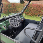 Green tractor cab interior featuring steering wheel, dashboard, and pedals; red hedge visible through the windows at the back.