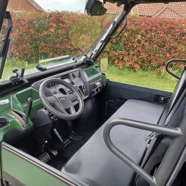 Green tractor cab interior featuring steering wheel, dashboard, and pedals; red hedge visible through the windows at the back.