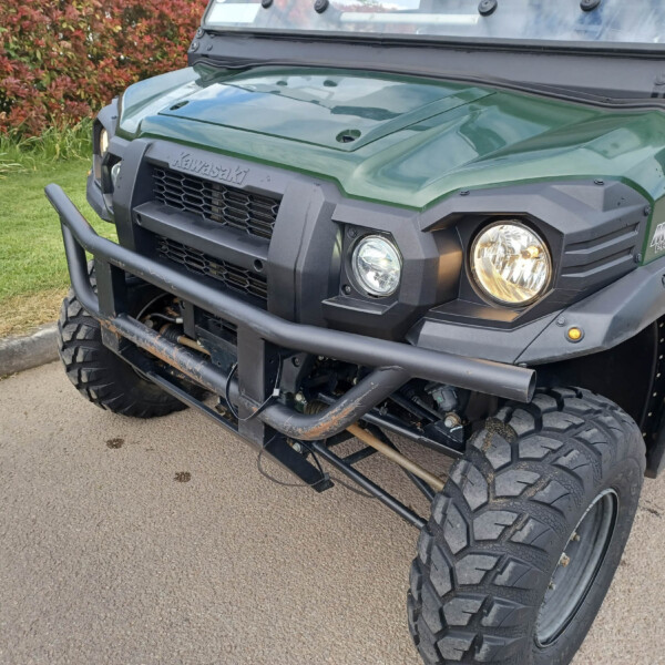Front view of a green Kawasaki off-road utility vehicle with round headlights and a rugged bumper, parked on a paved path with red shrubs in the background.
