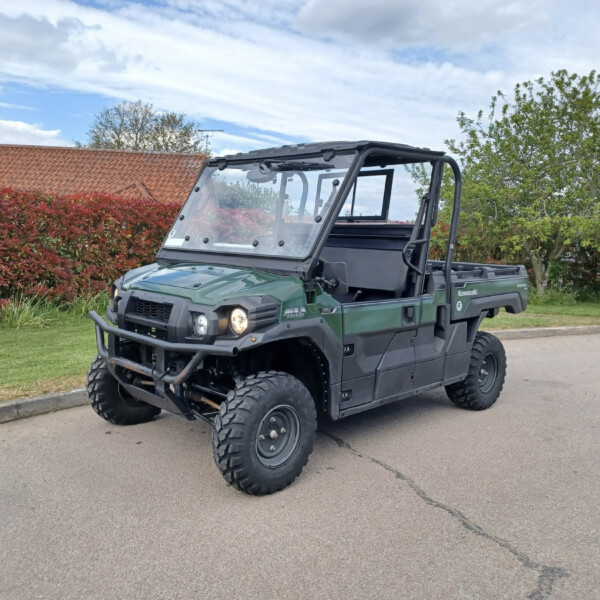 Front three-quarter view of a green Kawasaki UTV with a roll cage, parked in a suburban lot beside red hedges and a cloudy blue sky.