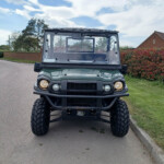 Front view of a green Kawasaki off-road utility vehicle with large tires, centered on a paved road and a clear windshield.