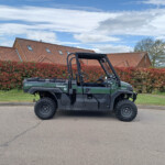 Green Kawasaki diesel 4x4 off-road utility vehicle with an open cabin parked on a street; red hedges and brick houses in the background.