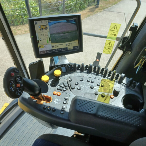 Inside the cab of a heavy machine showing a large monitor with camera view, gauges, and controls, plus joysticks and yellow levers in the foreground.