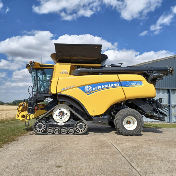 Yellow New Holland combine harvester with tracked front wheels parked beside a gray corrugated building on a sunny day.
