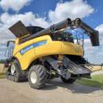 Yellow New Holland combine harvester with extended grain auger in a field under a blue sky with white clouds; large track tires visible on the front.