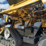 Close-up of a yellow agricultural combine harvester showing internal belts, gears and hydraulic hoses under the body.
