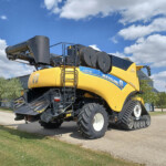 Yellow New Holland combine harvester with a raised header and crawler-style tracks parked on a paved lot under a blue sky with clouds.