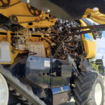 Close-up of a yellow construction machine's interior, showing belts, gears, hydraulic hoses and wiring amid exposed components and a large tire nearby.