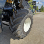 Close-up of a large tractor rear tire with a dark blue body and exposed hydraulic lines, parked on a street with a flag and buildings in the background.]","Close-up of a tractor’s oversized tire and machinery, showing hydraulic hoses and metal steps beside a paved road.