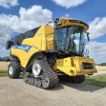 Yellow New Holland combine harvester with tracked wheels parked on a road under a blue sky with clouds.