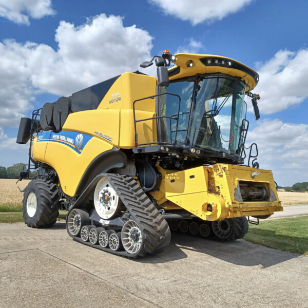 Yellow New Holland combine harvester with tracked wheels parked on a road under a blue sky with clouds.