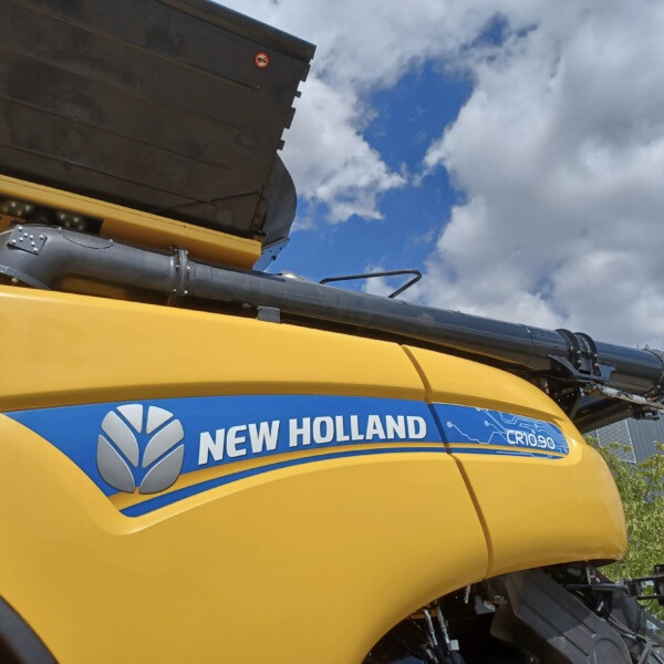 Close-up of a bright yellow New Holland agricultural machine with a blue decal and logo visible on the side, against a cloudy sky backdrop.