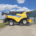 Yellow New Holland CR10.90 combine harvester with tracked rear wheels, parked beside a gray industrial building under a blue, cloudy sky.