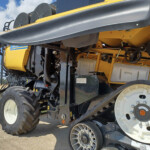 Close-up of a yellow agricultural harvester showing large black tires, gears, and track components against a rural field and blue sky.