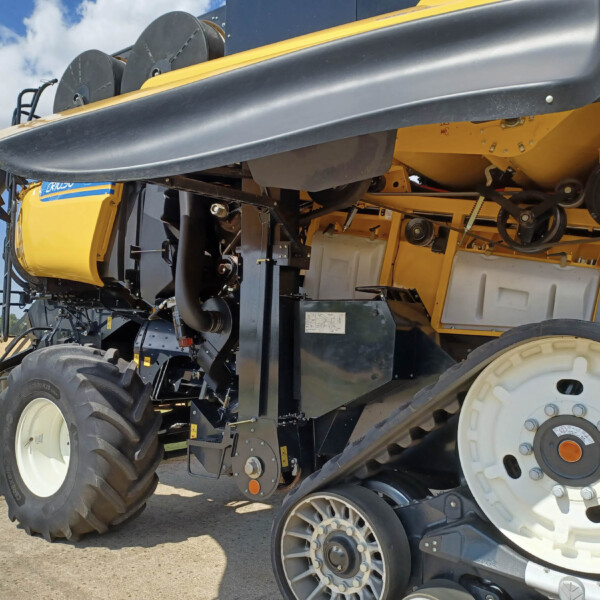 Close-up of a yellow agricultural harvester showing large black tires, gears, and track components against a rural field and blue sky.