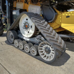 Close-up of a yellow tracked agricultural machine showing large rubber tracks and white drive wheels side view.