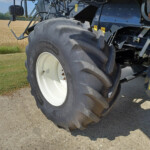 Close-up of a large tractor rear tire and white wheel, with blue machinery and yellow warning stickers, on a gravel path beside a wheat field.