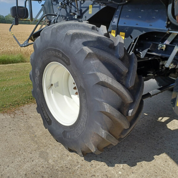 Close-up of a large tractor rear tire and white wheel, with blue machinery and yellow warning stickers, on a gravel path beside a wheat field.
