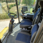 Interior view of a heavy machinery cab with steering wheel, joystick, and control panel; outdoor site visible through windshield.