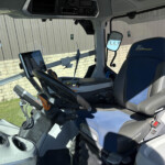 Interior of a heavy vehicle cab with a blue seat labeled 'Blue Power,' a mounted tablet, and a steering wheel visible at the front, with a gray industrial wall seen through the window outside.