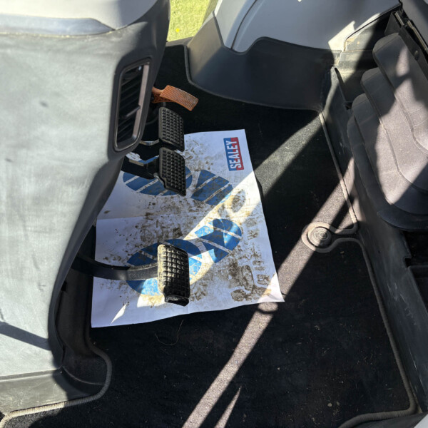 Driver-side car footwell showing accelerator and brake pedals, a black textured pedal cover, and a muddy paper mat with blue and red Sealey logo partially visible in bright sunlight.