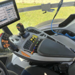 Tractor cab interior with a large touchscreen display showing engine data and controls, plus a joystick with orange buttons nearby.