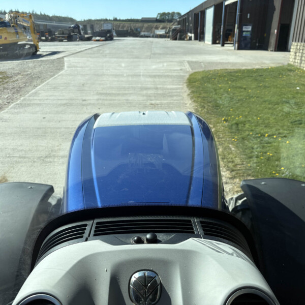 View from inside a vehicle looking over a blue hood at a construction yard: a yellow Komatsu excavator on the left, wide concrete lot, and large warehouse buildings on the right under a sunny sky.
