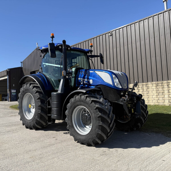 Blue New Holland tractor with large tires parked in an outdoor industrial yard beside a corrugated metal building under a clear blue sky, with orange warning lights on top of the cab.