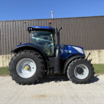 Front-left view of a blue agricultural tractor parked along a brown corrugated metal wall under a clear blue sky, with a sign on the right edge.