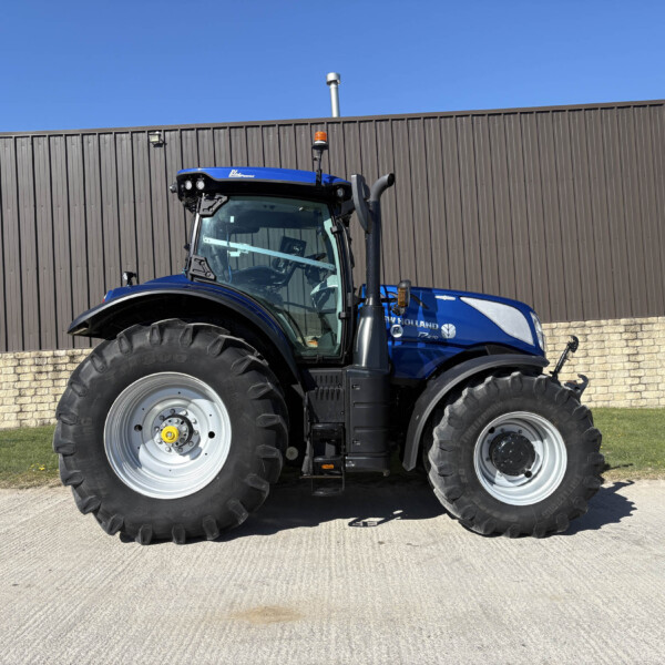 Front-left view of a blue agricultural tractor parked along a brown corrugated metal wall under a clear blue sky, with a sign on the right edge.