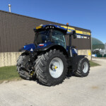 Blue tractor with oversized tires parked in front of a brown industrial building under a clear blue sky.